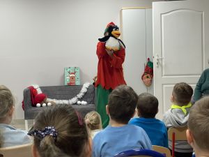 Dzieci siedzą na krzesełkach i oglądają Panią w stroju Elfa która trzyma pacynkę pingwina