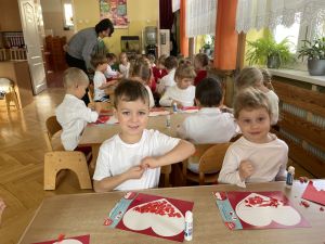 Dzieci siedzą przy stolikach i wykonują pracę plastyczną. Wyklejają serduszka czerwoną bibułą