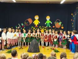 Dzieci siedzą w półkolu na krzesełkach. Ubrane są w ludowe stroje. Na środku stoi dwóch chłopców i jedna dziewczynka i mówią do mikrofonu