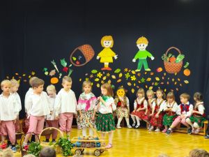 Dzieci siedzą w półkolu na krzesełkach. Ubrane są w ludowe stroje. Na środku stoi 6 chłopców i 1 dziewvzynka