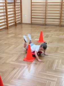 Dziewczynka jedzie slalomem między pachołkami na deskorolce gimnastycznej leżąc na brzugu