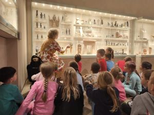 Dzieci siedzą na podłodze i słuchają edukatora muzealnego.  Pani opowiada o faraonach