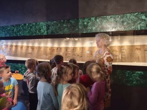 Dzieci siedzą na podłodze i słuchają edukatora muzealnego.  W witrynie widać papirus