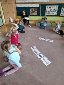 Dzieci siedzą na dywanie, przed nimi leżą obrazki z różnymi zwierzętami. Pod tablicą siedzi nauczycielka 