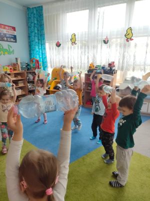 Dzieci stoją na dywanie i trzymają butelki uniesione nad głowami