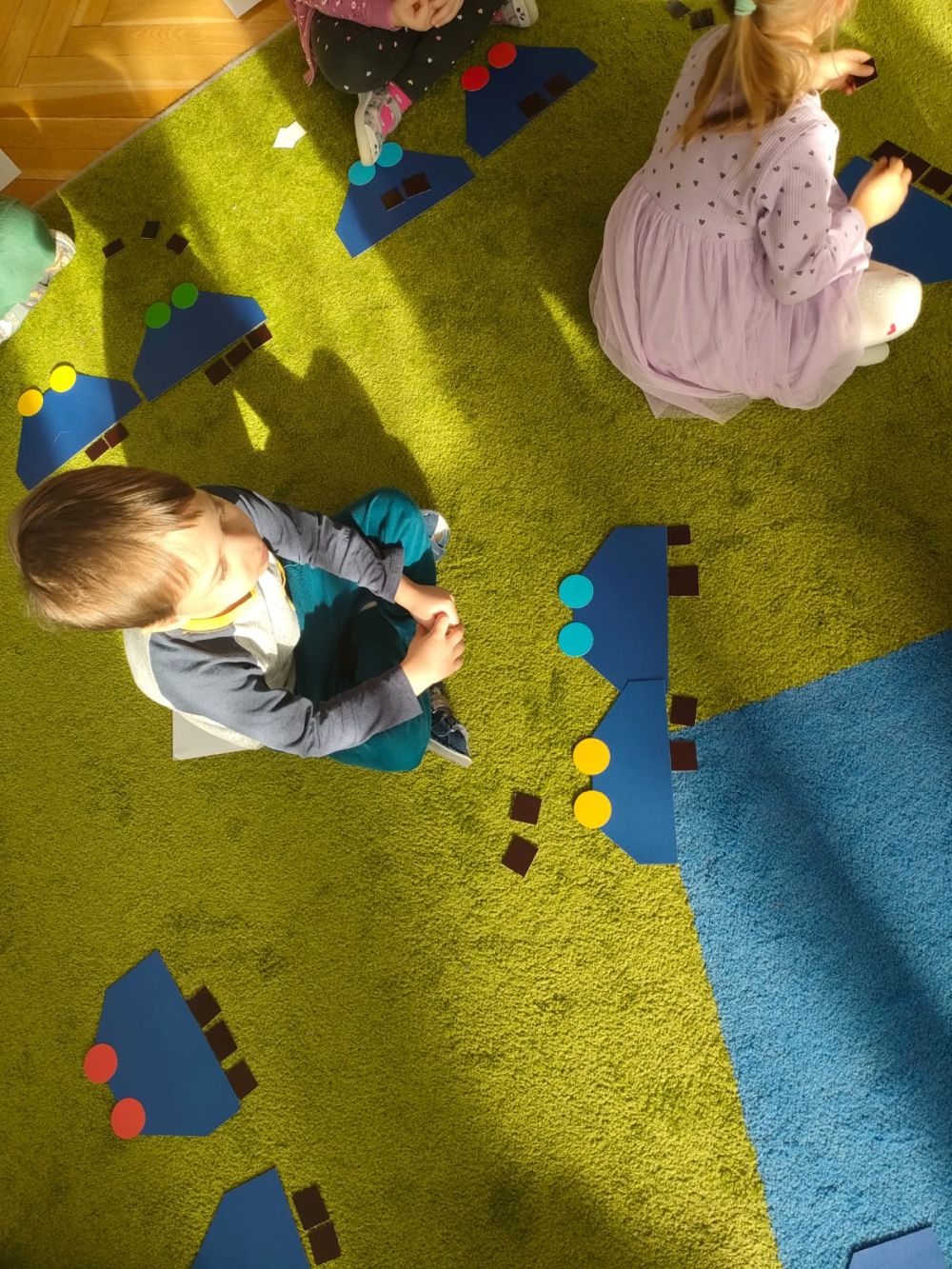 Dzieci siedzą na dywanie. Przed nimi leżą papierowe wagoniki i czarne kwadraciki
