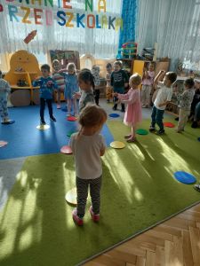 Dzieci tańczą na dywanie, na którym rozłożone są kolorowe dyski.