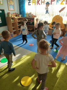 Dzieci tańczą na dywanie, na którym rozłożone są kolorowe dyski.