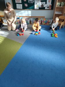 Na dywanie siedzi nauczycielka i  dzieci. Troje dzieci buduje z klocków domki