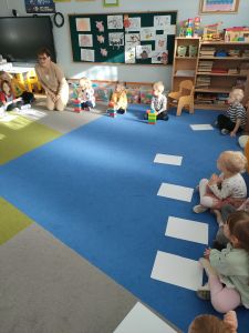 Na dywanie siedzi nauczycielka i  dzieci. Troje dzieci buduje z klocków domki