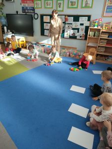 Na dywanie siedzi nauczycielka i  dzieci. Troje dzieci buduje z klocków domki