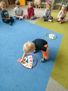 Dzieci siedzą w kole na dywanie. Chłopiec wybiera obrazek z parasolką.