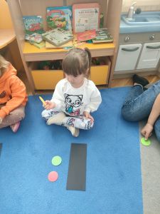 Dziewczynka siedzi na dywanie a przed nią leży czarna prostokątna kartka i kółka w kolorze zielonym i czerwonym. Dziewczynka trzyma żółte kółko