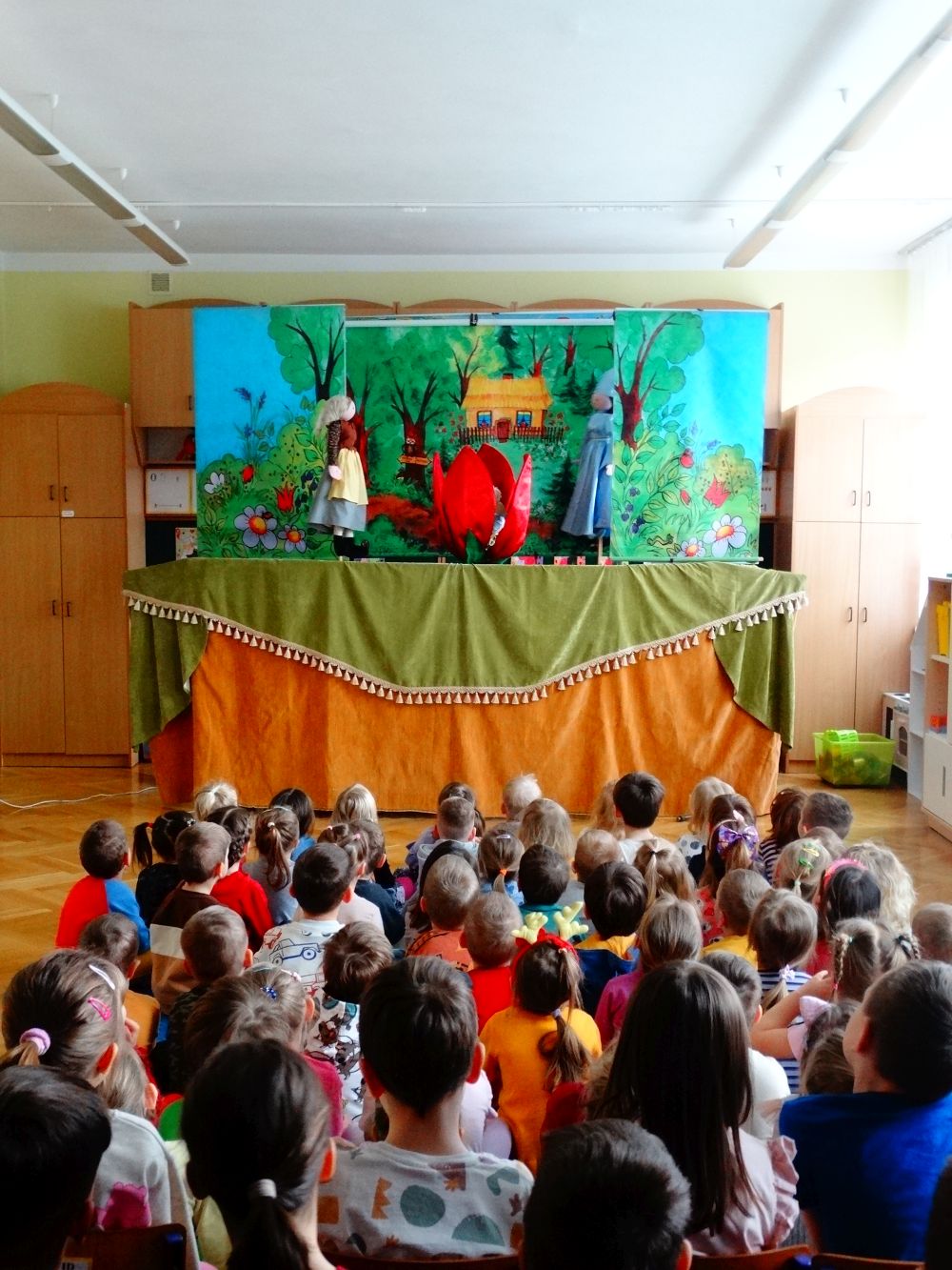 Dzieci siedzą na dywanie. Patrzą na teatrzyk kukiełkowy.