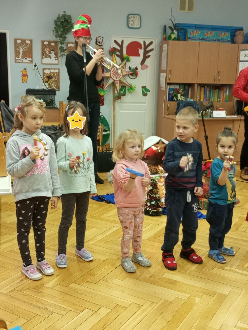 Dzieci stoją na scenie i grają na dzwoneczkach. W tyle  Pani gra na trąbce a pan na gitarze