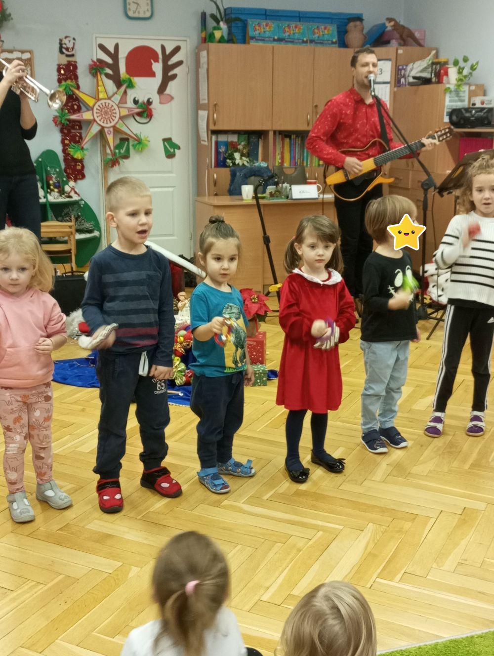 Dzieci stoją na scenie i grają na dzwoneczkach. W tyle  Pani gra na trąbce a pan na gitarze