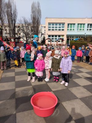 dzieci z wszystkich grup stoją na placu zabaw, 5 dziewczynek stoi na środku a jedna z nich trzyma marzannę