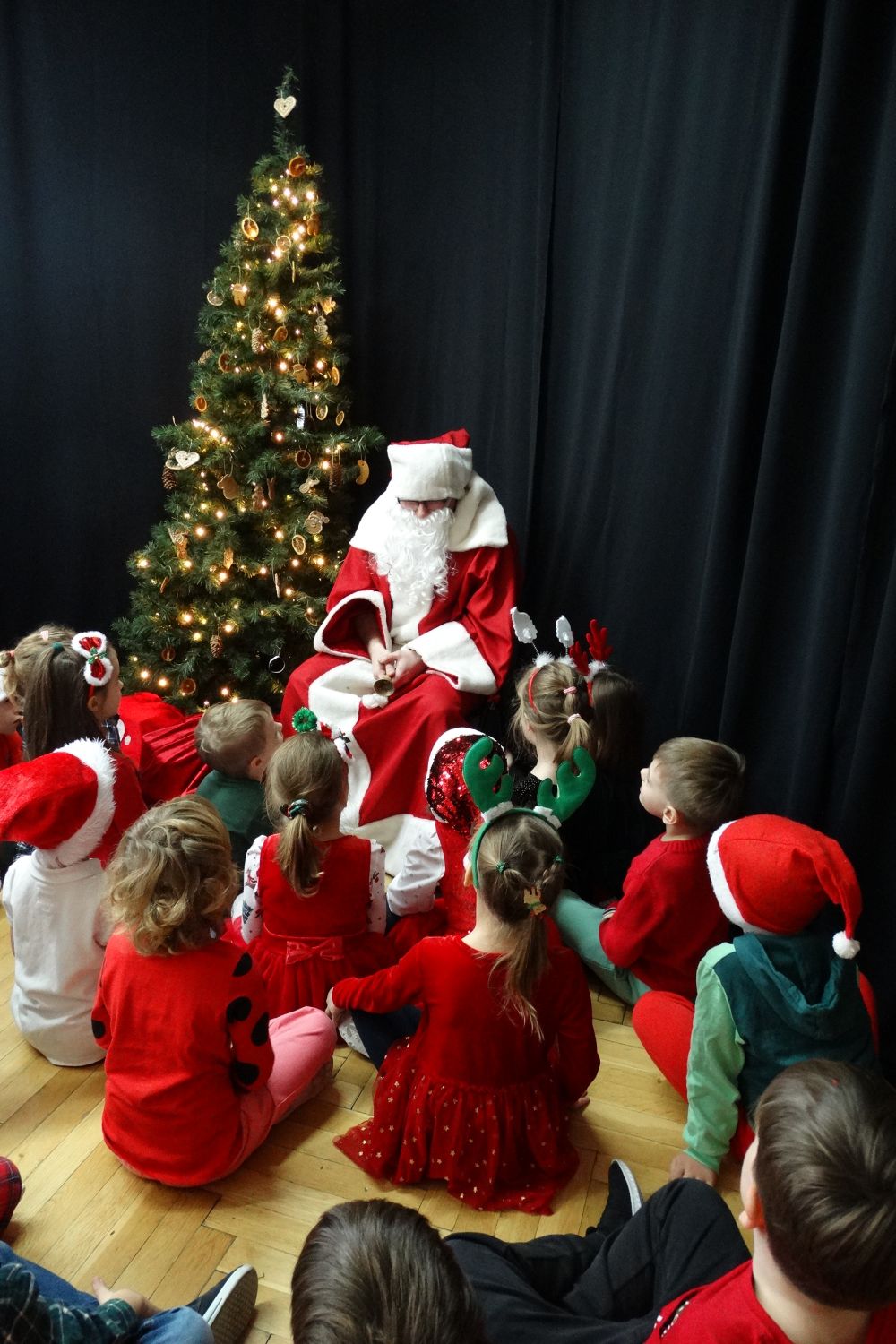 Dzieci z grupy 5 siedzą na podłodze i patrzą w stronę świętego mikołaja. Mikołaj siedzi na krześle obok przystrojonej choinki