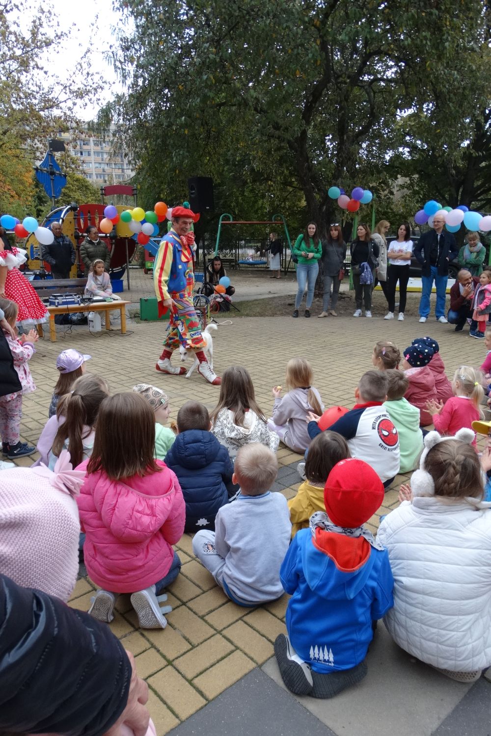 Dzieci są na placu zabaw i patrzą na pana ubranego w kolorowy strój który pokazuje sztuczki z psem