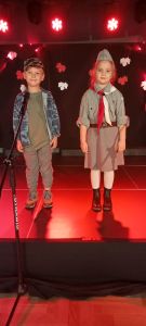 Dzieci stoją na scenie. Chłopiec jest ubrany w strój wojskowy, dziewczynka ubrana jest w mundur harcerski