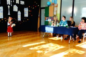 Dziewczynka ubrana w stój krakowski stoi na środku sceny i mówi do mikrofonu.  W tle dekoracja przedstawiające białe budynki. Z prawej strony siedzi komisja konkursowa