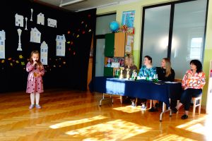 Dziewczynka stoi na środku sceny i mówi do mikrofonu. W ręce trzyma piernikowe serduszko. W tle dekoracja przedstawiające białe budynki. Z prawej strony siedzi komisja konkursowa