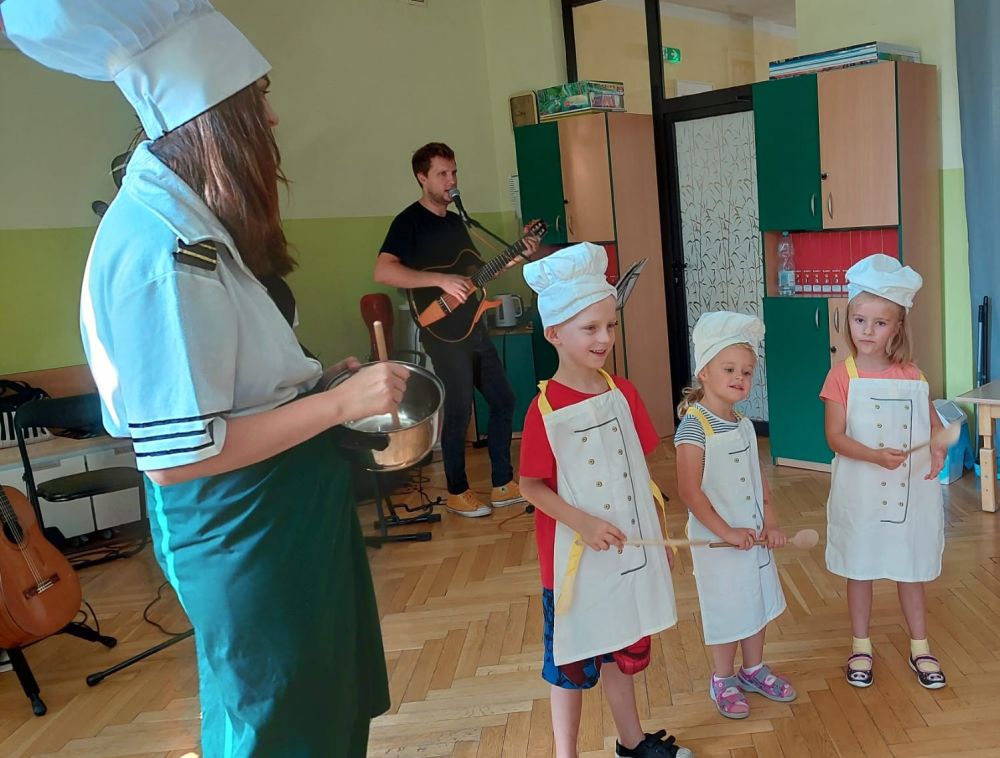 Pan gra na gitarze. Pani ma czapkę kucharza i trzyma garnek. Troje dzieci stoi w strojach kucharzy z drewnianymi łyżkami w rękach