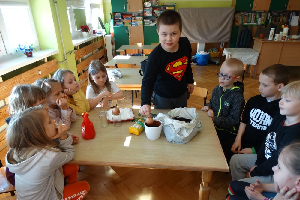 Na zdjęciu widać 6 dziewczynek i 4 chłopców z grupy 5. Dzieci siedzą dookoła stolika, na którym stoi konewka, doniczka wypełniona ziemią, tacka z watą, torba z ziemią. Jeden z chłopców wkłada cebulę do ziemi.