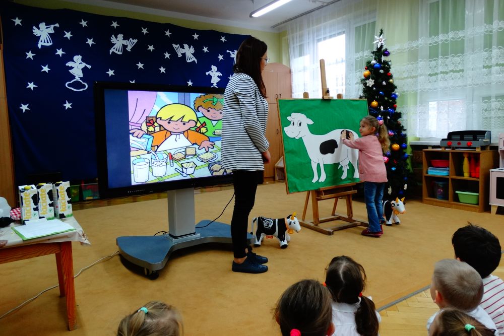 Na zdjęciu dzieci z całego przedszkola siedzą tyłem, patrzą jak dziewczynka przyczepia łaty na papierowej krowie