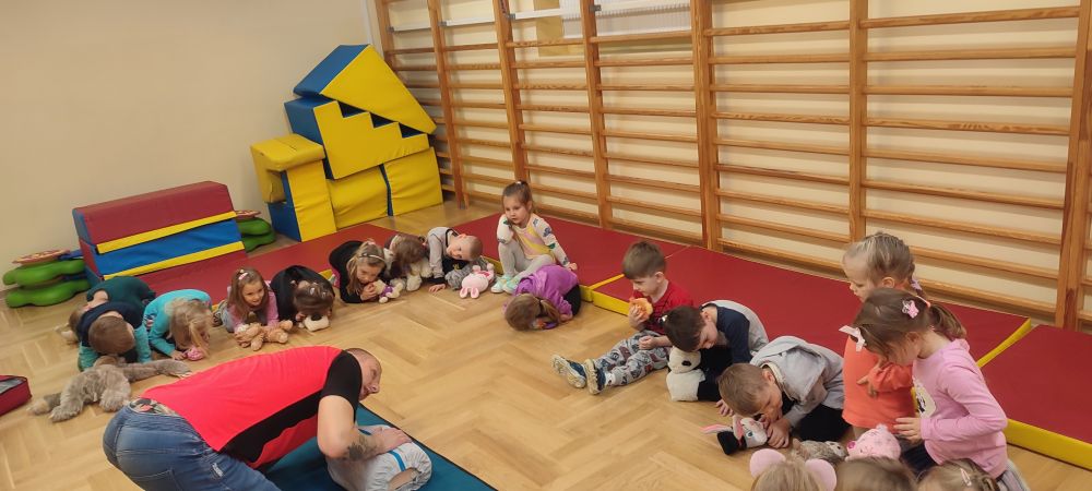 Ratownik medyczny jest schylony nad fantomem nasłuchując oddechu, dzieci naśladują ratownika nachylając się policzkami nad swoimi pluszakami.