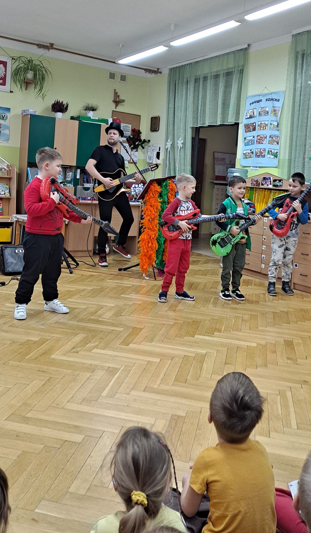 Kilku chłopców stoi na scenie i trzyma w rękach dmuchane gitary. Obok stoi Pan z melonikiem gra na gitarze elektrycznej. Inne dzieci siedzą na dywanie