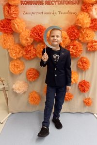 Chłopiec stoi a w tle dekoracja kwiatowa z napisem Konkurs recytatorski