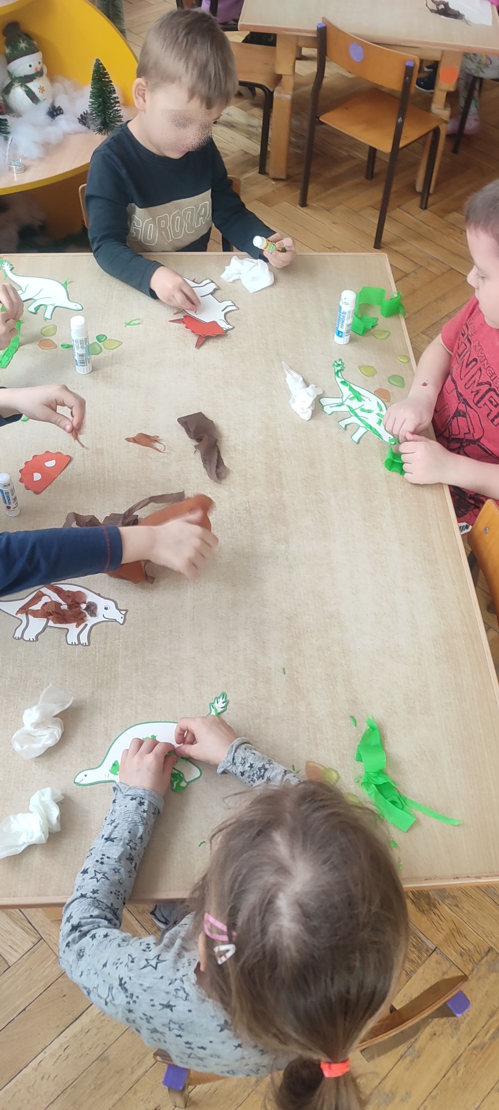 Chłopcy siedzą przy stoliku i wyklejają bibułą dinozaury