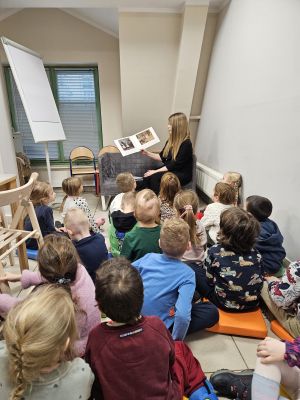 Dzieci siedzą na poduszkach i słuchają ksiąąki czytanej przez Panią z biblioteki