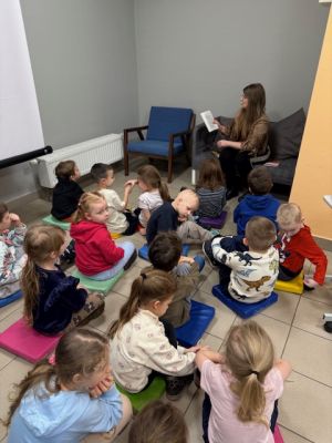 Dzieci siedzą na poduszkach i słuchają książki czytanej przez Panią z biblioteki