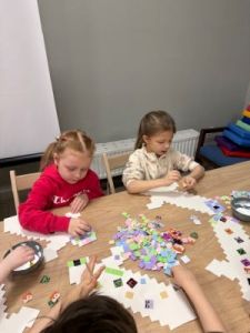 Dwie dziewczynki siedzą przy stole i wyklejają kwadracikami kartonowy miecz dobra