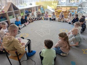 Chłopiec siedzi na krześle i czyta książkę. Dzieci siedzą w kole na dywanie