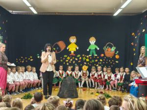 Dzieci siedzą w półkolu na krzesełkach. Ubrane są w ludowe stroje. Pani dyrektor stoi na środku i wita zebranych gości.