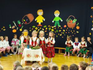 Dzieci siedzą w półkolu na krzesełkach. Ubrane są w ludowe stroje. Na środku stoją 4 dziewczynki i mówią do mikrofonu