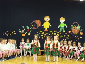 Dzieci siedzą w półkolu na krzesełkach. Ubrane są w ludowe stroje. Na środku stoją 3 dziewczynki i mówią do mikrofonu