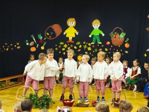 Dzieci siedzą w półkolu na krzesełkach. Ubrane są w ludowe stroje. Na środku stoi 6 chłopców ubranych w stroje ludowe