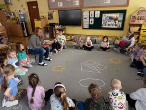 Mama Wiktorii czyta dzieciom książkę. Dzieci siedzą w kole na dywanie