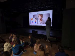 Dzieci siedzą i patrzą na wyświetlony na ekranie obrazek zezłoszczonego chłopca