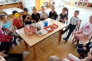 Dzieci siedzą dookoła stolika na którym leżą przygotowane rzeczy do siania i sadzenia 