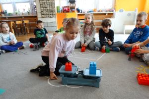 Dzieci siedzą na dywanie. Dziewczynka patrzy na klocki postawione na wadze szalkowej