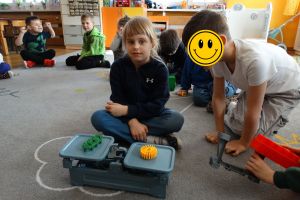 Dzieci siedzą na dywanie. Dziewczynka patrzy na klocki postawione na wadze szalkowej