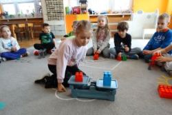 Dzieci siedzą na dywanie. Dziewczynka patrzy na klocki postawione na wadze szalkowej