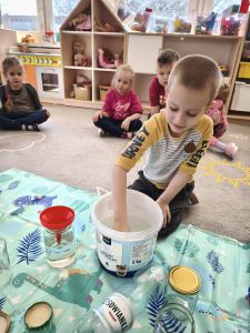 Chłopiec nabiera wodą do szklanki z wiaderka. Dzieci siedzą na dywanie
