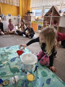 Dzieci siedzą na dywanie. Dziewczynka wlewa wodę do słoika