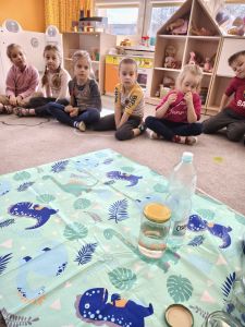 Dzieci siedzą na dywanie i porównują ilość wody w butelce i słoiku